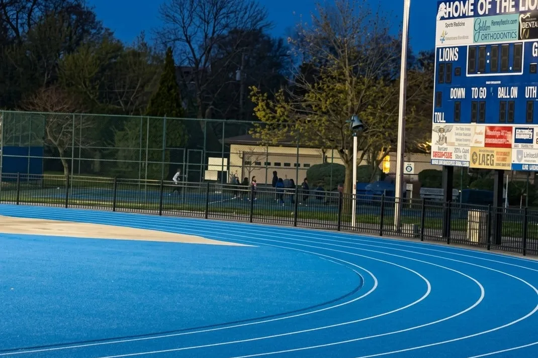 The resurfaced Bexley blue running track curving past the stadium scoreboard at dusk
